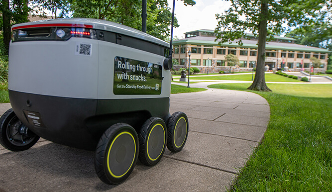 A small delivery robot on a campus path, with the text "Rolling through with snacks." A modern building and lush greenery are in the background.